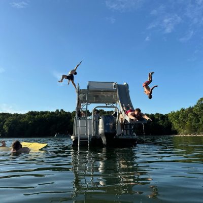 People jumping into a lake from a boat with a slide on a sunny day.
