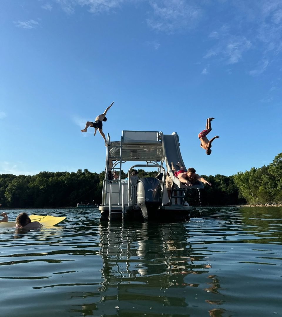 People jumping into a lake from a boat with a slide on a sunny day.